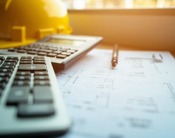 Top-down view of a construction company office desk with calculator, invoice papers and a yellow hard hat near a window with warm light